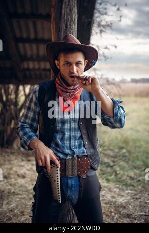 Brutal cowboy smokes a cigar, texas ranch on background, western. Vintage male person with gun on farm, wild west culture Stock Photo