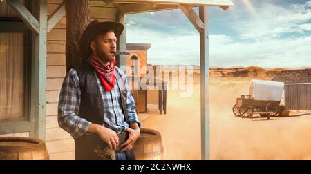 Brutal cowboy smokes a cigar at the saloon entrance, desert valley on background, western. Vintage male person with gun in country, wild west culture Stock Photo