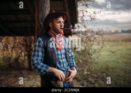 Brutal cowboy smokes a cigar, texas ranch on background, western. Vintage male person with gun on farm, wild west Stock Photo