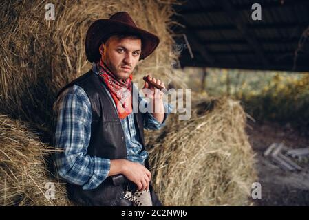 Brutal cowboy smokes a cigar, haystack on background, western. Vintage male person with gun on farm, wild west culture Stock Photo