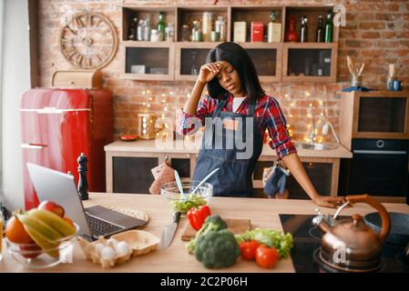 Sleepy black woman in apron cooking healthy breakfast on the kitchen. African female person preparing vegetable salad at home Stock Photo