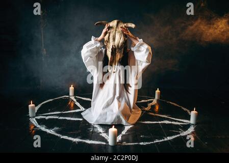 Young woman in white shirt holds skull of the animal in hands, pentagram circle with candles, smoke all around. Dark magic ritual, occultism
