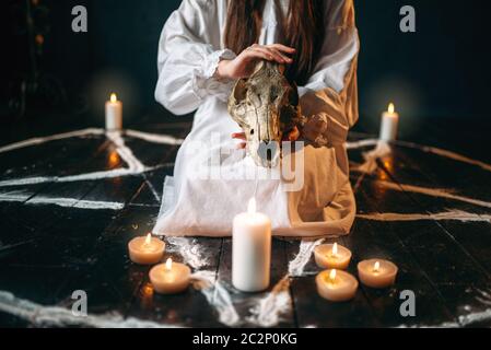 Female person in white shirt holds animal skull in hands, pentagram circle with candles. Dark magic ritual, occultism