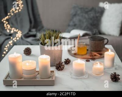 Cozy home, hygge, cosiness concept - burning white fragrance candles on white marble table near sofa with pillows and plaid. Winter decor, hot tea cup Stock Photo