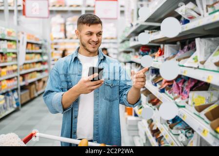 Man with cart makes a purchase by the list on phone in supermarket. Male customer in shop, husband with trolley choosing consumer goods, family shoppi Stock Photo