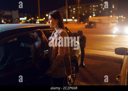 Female driver at the car, robber sneaking up behind, criminal, stealing. Hijacker victim in vehicle on parking. Auto robbery, automobile crime, hijack Stock Photo