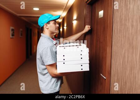 Pizza delivery boy knocking on the door of the customer, delivering service. Courier from pizzeria holds cardboard packages indoors Stock Photo