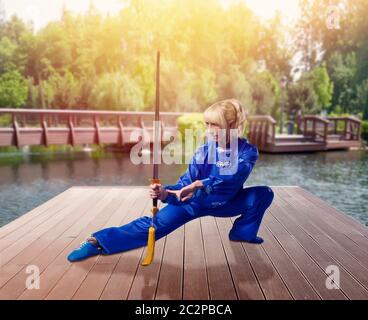 Female wushu fighter with blade against lake and green nature, martial arts. Woman in blue cloth on outdoor fight training Stock Photo