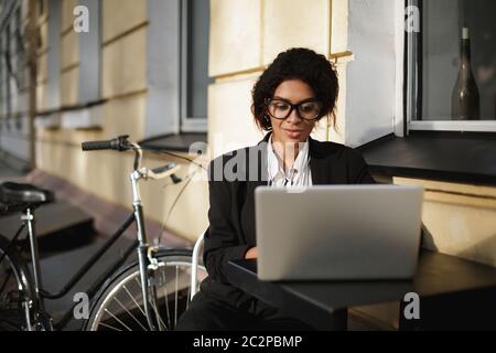 Portrait of African American girl in glasses sitting at the table of cafe and working on her laptop. Smiling lady with dark curly hair in black jacket Stock Photo