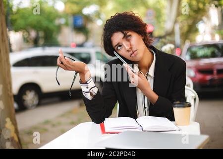 Portrait of African American girl sitting at the table with glasses in hand and dreamily looking up while talking on her cellphone. Lady with dark cur Stock Photo