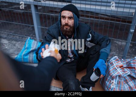 Male person gives food to bearded dirty beggar on city street. Poverty is a social problem, homelessness and loneliness, alcoholism and  drunk addicti Stock Photo