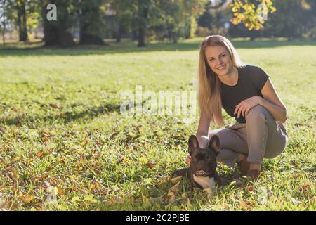 Smiling long haired young girl posing with cute French Bulldog in autumn park looking at the camera. Stock Photo