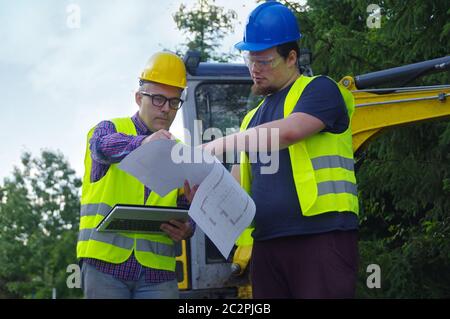 Engineer and worker on construction site with excavator in the background. A professional team working at the streets building. Two men in helmets tal Stock Photo