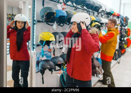 Young woman trying on helmet for ski or snowboarding at the mirror, side view, sports shop. Winter season extreme lifestyle, active leisure store, buy Stock Photo