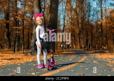 Cute adorable little caucasian school girl portrait having fun riding pink kick scooter bike in golden autumn city park or forest outside. Happy Stock Photo