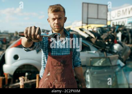 Male repairman shows wrench on car scrapyard. Auto scrap, vehicle junk, automobile garbage. Abandoned, damaged and crushed transport, junkyard Stock Photo