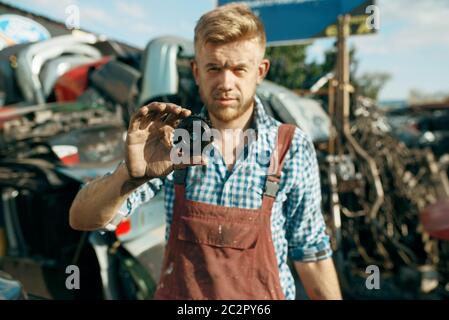 Male mechanic shows gear car on junkyard. Auto scrap, vehicle junk, automobile garbage. Abandoned, damaged and crushed transport, scrapyard Stock Photo