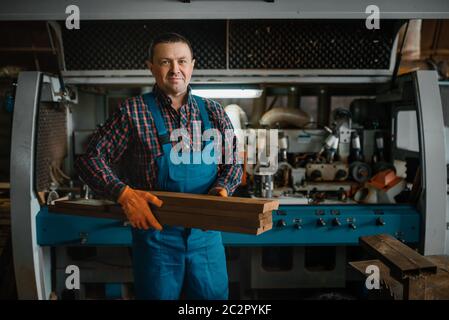 Carpenter in uniform holds boards, woodworking machine on background, lumber industry, carpentry. Wood processing on factory, forest sawing in lumbery Stock Photo