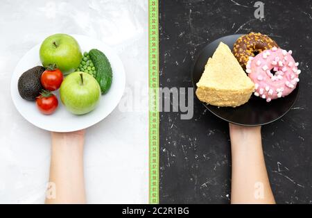 Female hands holding white plate with vegetables and fruits and black plate with donuts and a cake on contrast background divided with a measuring tap Stock Photo