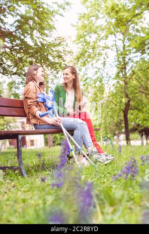 Two women, one healthy and one with a sprained foot, on a bench in the park Stock Photo