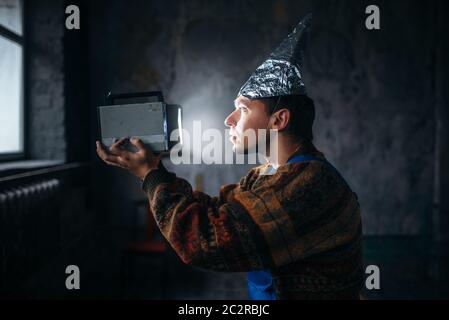 Paranoid man in tinfoil cap watch TV, mind protection from telepathy, paranoia concept. UFO phobia Stock Photo