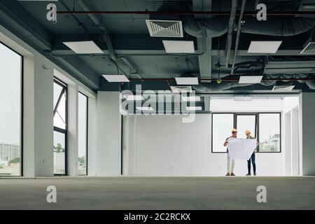 Two workers wearing hardhats standing in big empty hall of new building looking at construction plan, extreme long shot Stock Photo