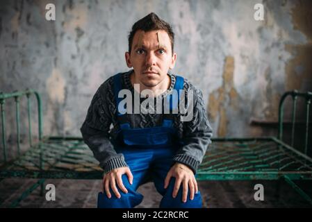 Mad man sitting on a rusty bed, opened eyelids sealed with plaster, depressed psycho patient. Mentally ill people concept, crazy human Stock Photo