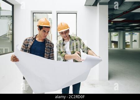 Two professional engineers wearing checked shirts and hardhats working on construction plan, horizontal medium long shot Stock Photo