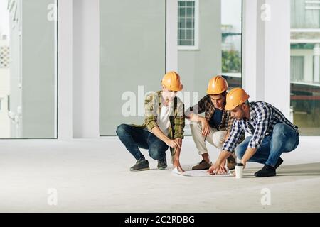 Group of three modern construction engineers wearing casual clothes and hardhats sitting on hunkers discussing building plan, copy space Stock Photo