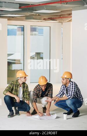 Vertical shot of three professional engineers sitting on hunkers in empty room of new building correcting construction plan Stock Photo