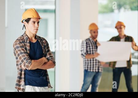 Portrait of professional architect wearing hardhat standing with arms crossed looking away, his colleagues working behind him Stock Photo