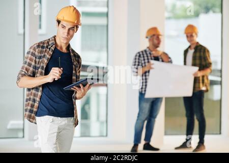 Professional architect with serious facial expression holding digital tablet computer looking away, his colleagues working behind him Stock Photo