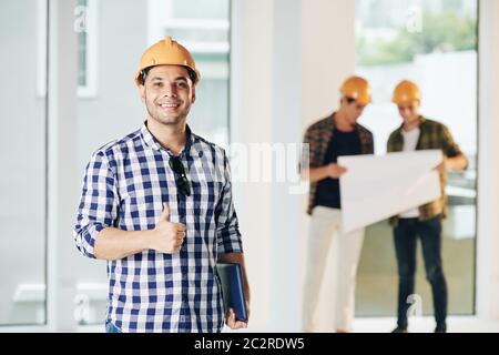 Medium portrait of construction engineer wearing casual shirt and hardhat looking at camera showing thumb up, his colleagues looking at building plan Stock Photo