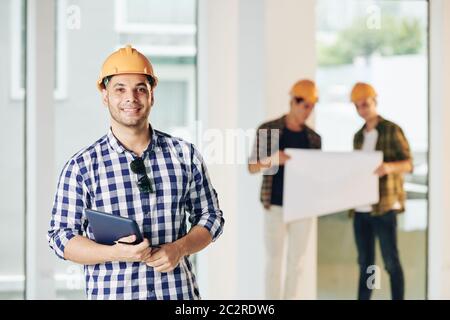 Medium portrait of professional engineer wearing casual shirt and hardhat holding tablet looking at camera, his colleagues looking at building plan Stock Photo