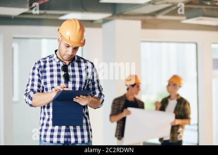 Portrait of professional engineer wearing casual shirt and hardhat using digital tablet for work, his colleagues discussing building plan Stock Photo