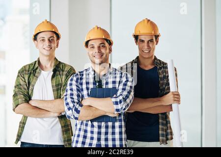 Horizontal medium shot of three professional engineers wearing checked shirts and hardhats standing together with arms crossed looking at camera smili Stock Photo