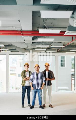 Vertical long shot of three professional engineers wearing checked shirts and hardhats standing together inside new building space Stock Photo