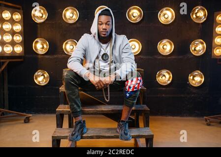 Black rapper in hoodie sitting on the steps, perfomance on stage with spotlights on background. Rap performer on scene with lights, underground music, Stock Photo