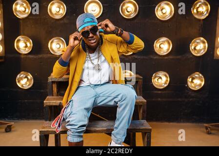 Black rapper in cap and sunglasses sitting on the steps, singer on stage with spotlights on background. Rap performer on scene with lights, undergroun Stock Photo