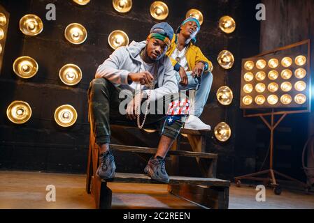 Two black rappers sitting on the steps, perfomance on stage with spotlights on background. Rap performers on scene with lights, underground music, urb Stock Photo