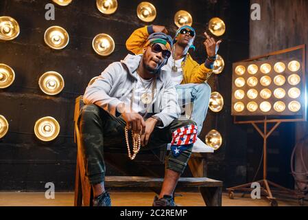 Two black rappers sitting on the steps, perfomance on stage with spotlights on background. Rap performers on scene with lights, underground music, urb Stock Photo