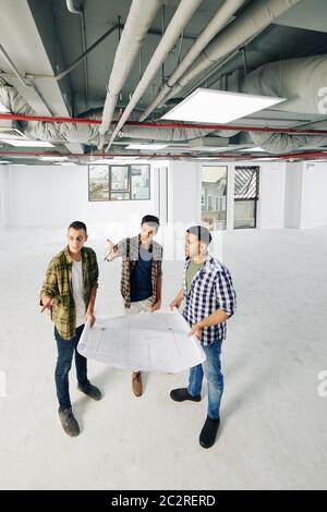 Group of professional engineers standing in empty spacious room holding building plan paper doing brainstorming, vertical high angle long shot, copy s Stock Photo