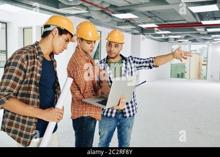 Three modern construction workers wearing hardhats searching additional information for current building project using laptop, copy space Stock Photo
