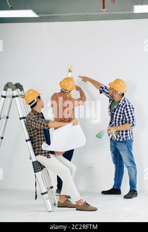 Modern manual workers wearing yellow hardhats checking unfinished room wall level, vertical shot Stock Photo