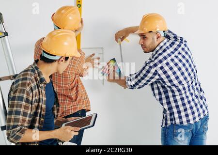 Group of three modern manual workers wearing checked shirts and yellow hardhats taking measurements for further work Stock Photo