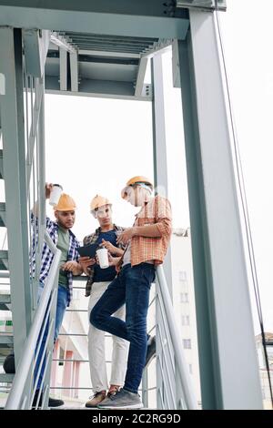 Vertical low angle shot of three modern professional construction engineers standing relaxed upstairs chatting during coffee break, copy space Stock Photo