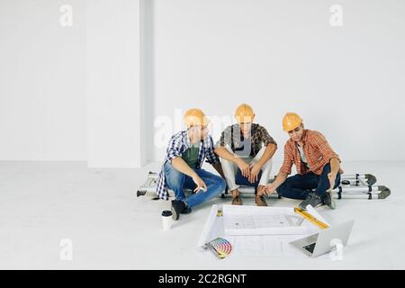 Middle Eastern men wearing casual clothes and hardhats sitting together in empty room with papers and laptop working on building plan Stock Photo