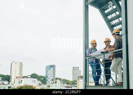 Group of three young construction engineers standing on fire-escape of new building holding paper plan and talking, copy space Stock Photo
