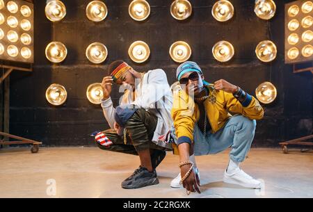 Two black rappers sitting on the floor, perfomance on stage with spotlights on background. Rap performers on scene with lights, underground music, urb Stock Photo