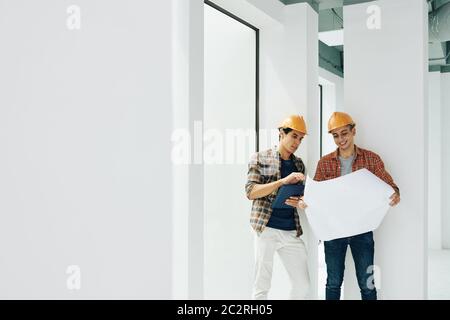 Two professional construction engineers standing together in unfinished room working with building plan paper, copy space Stock Photo